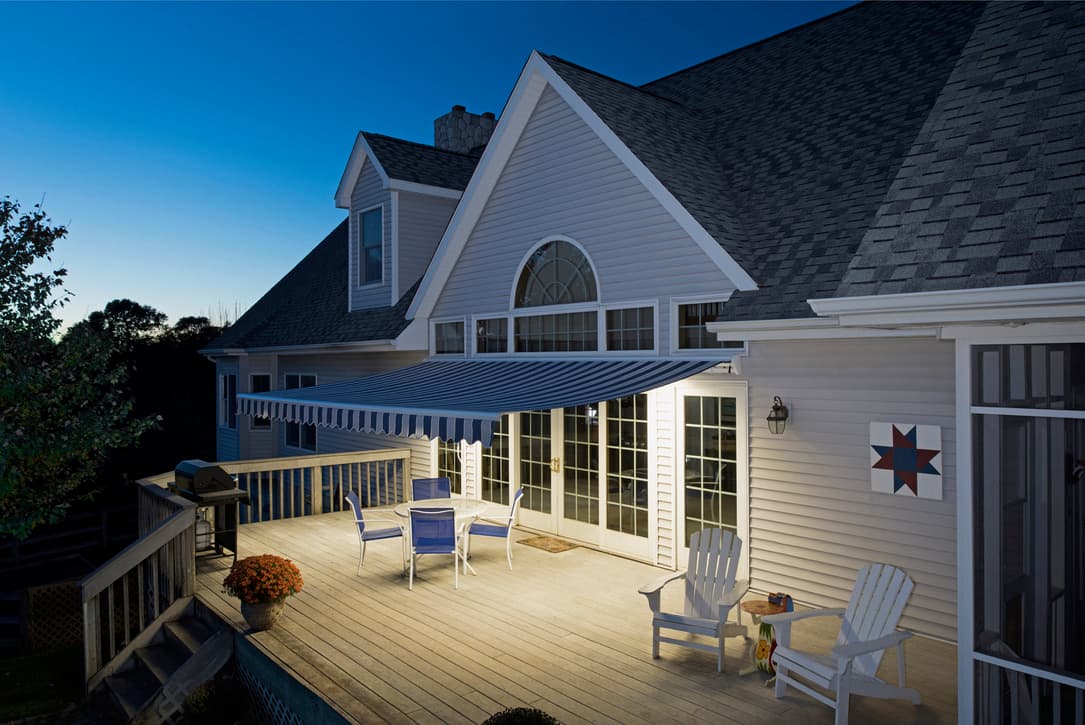 Blue-striped retractable awning extended over a backyard deck at dusk with lit interior